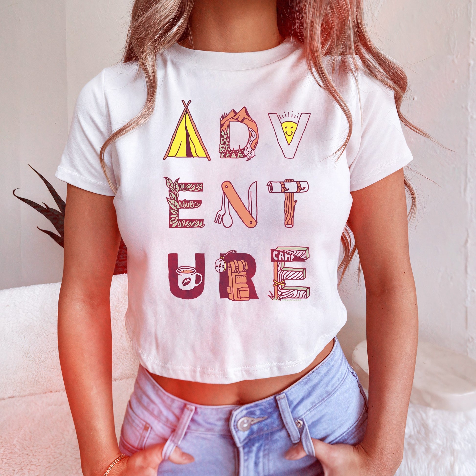 Adventure Life Women's Baby Tee features the word ADVENTURE styled with camping icons like a tent and backpack. This playful, cropped T-shirt embodies a spirited, outdoor-loving lifestyle.