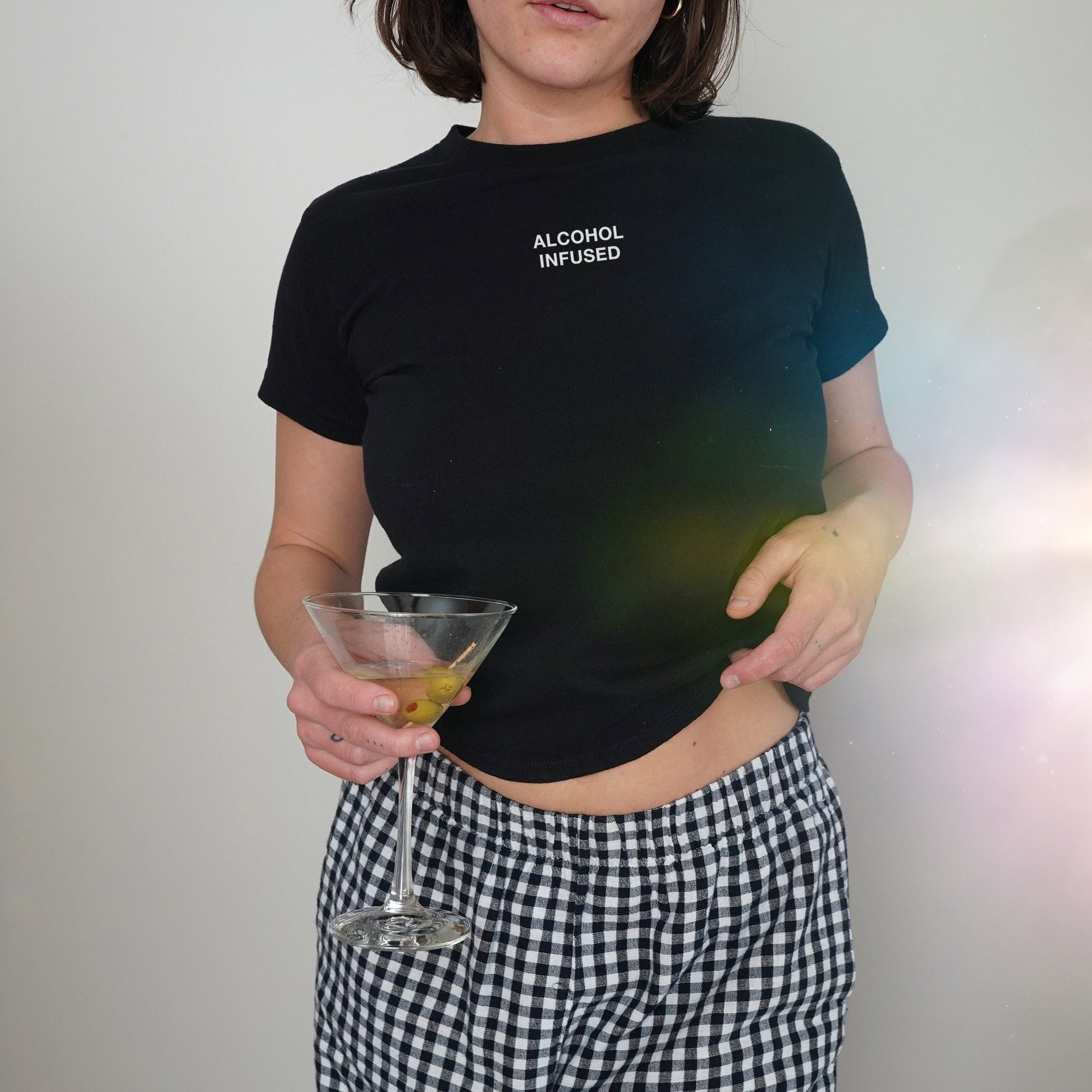 Alcohol Infused Women's Baby Tee: Black cropped T-shirt with 'ALCOHOL INFUSED' print, styled with gingham pants. Model holds martini glass with olives, capturing a bold, expressive vibe.