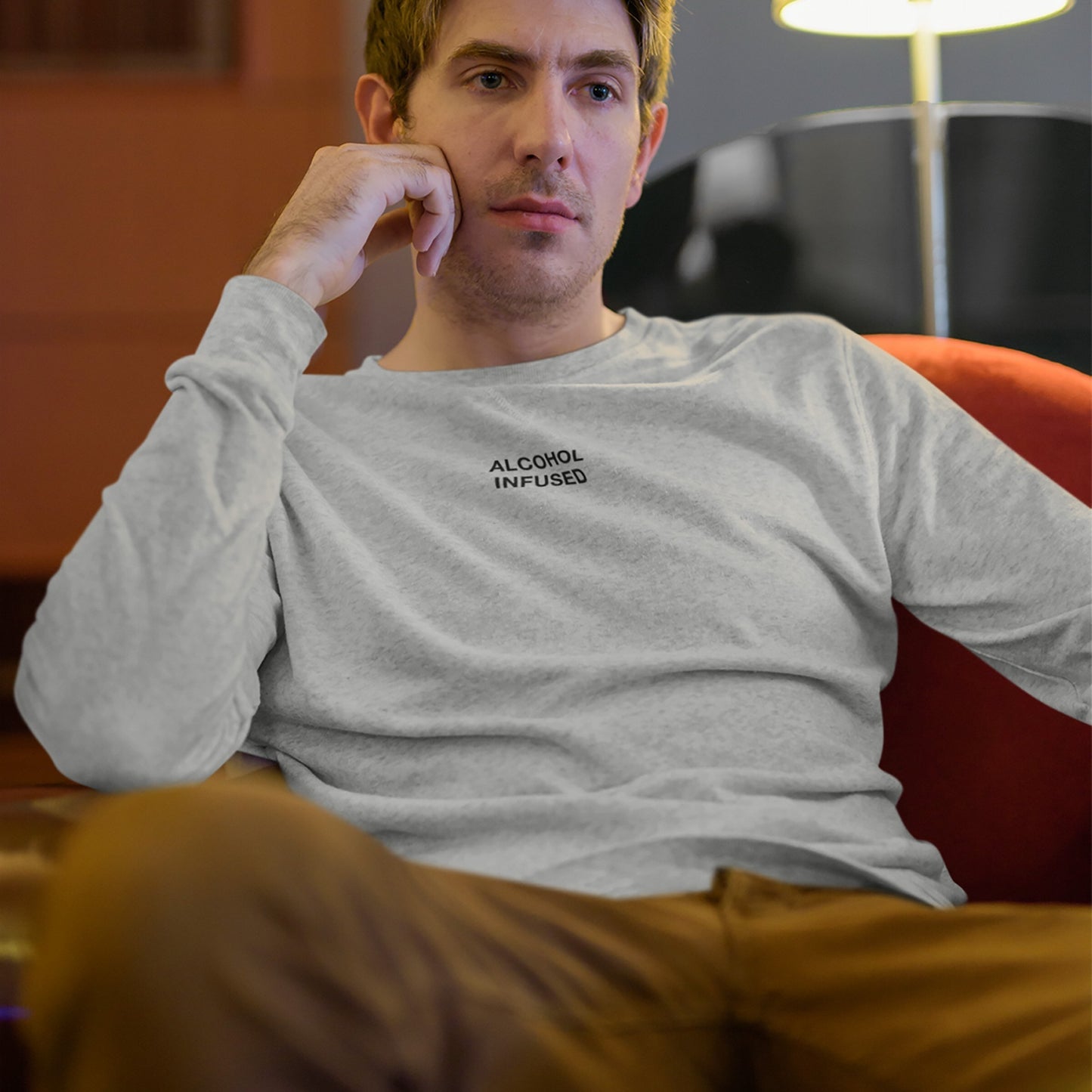 Young man reclines in a red armchair wearing the Alcohol Infused T-shirt, exuding a laid-back vibe amidst a casual indoor setting, embodying unapologetic individuality and subtle rebellion.