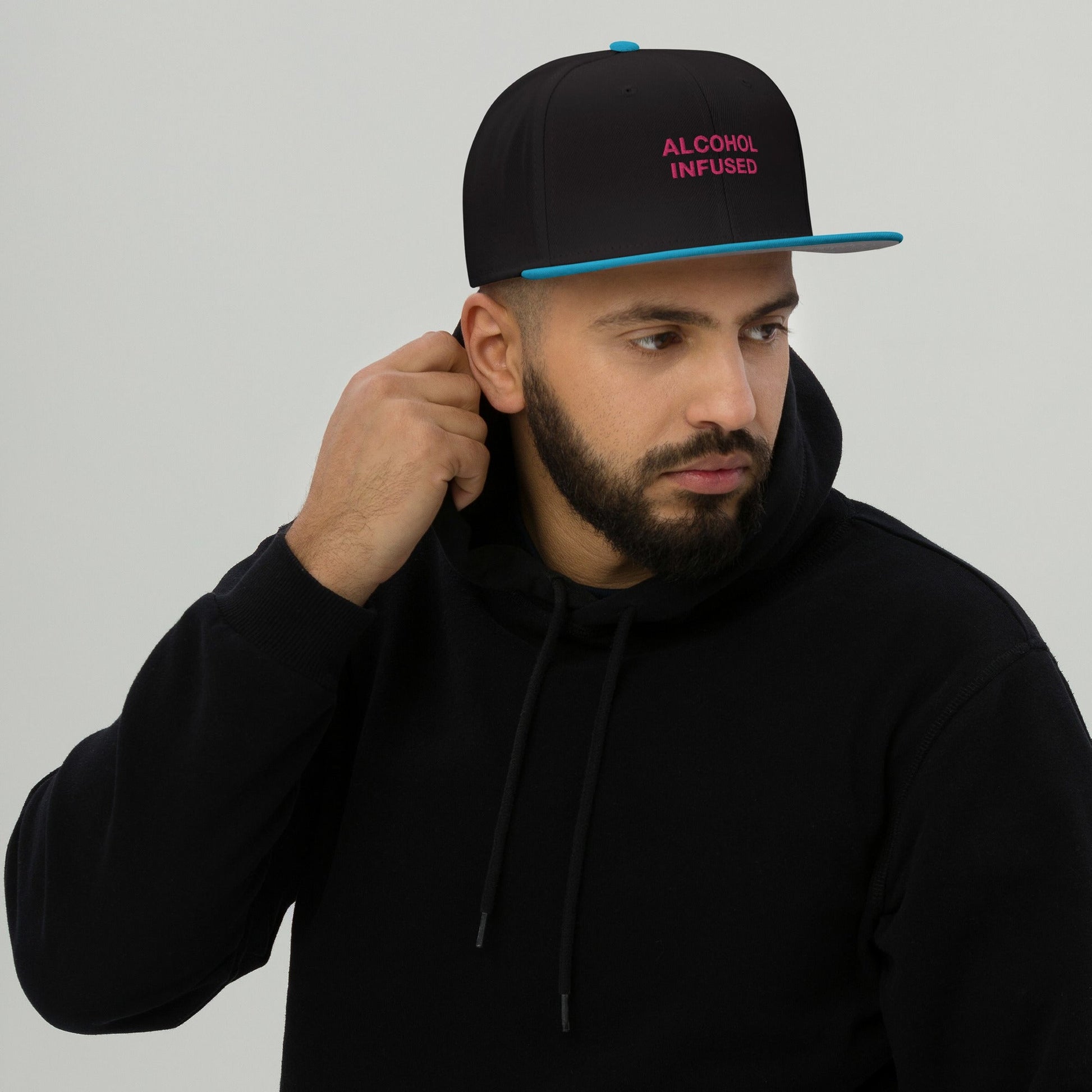 Young man models the Alcohol Infused Hat, a black snapback with teal accents and bold pink lettering, embodying edgy, rebellious style perfect for making bold statements.