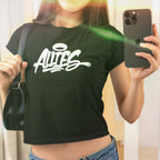 Young woman in mirror selfie wearing the Allies Handstyle Women's Baby Tee, showcasing the graffiti-style ALLIES print with halo. She's holding a dark shoulder bag and smartphone.