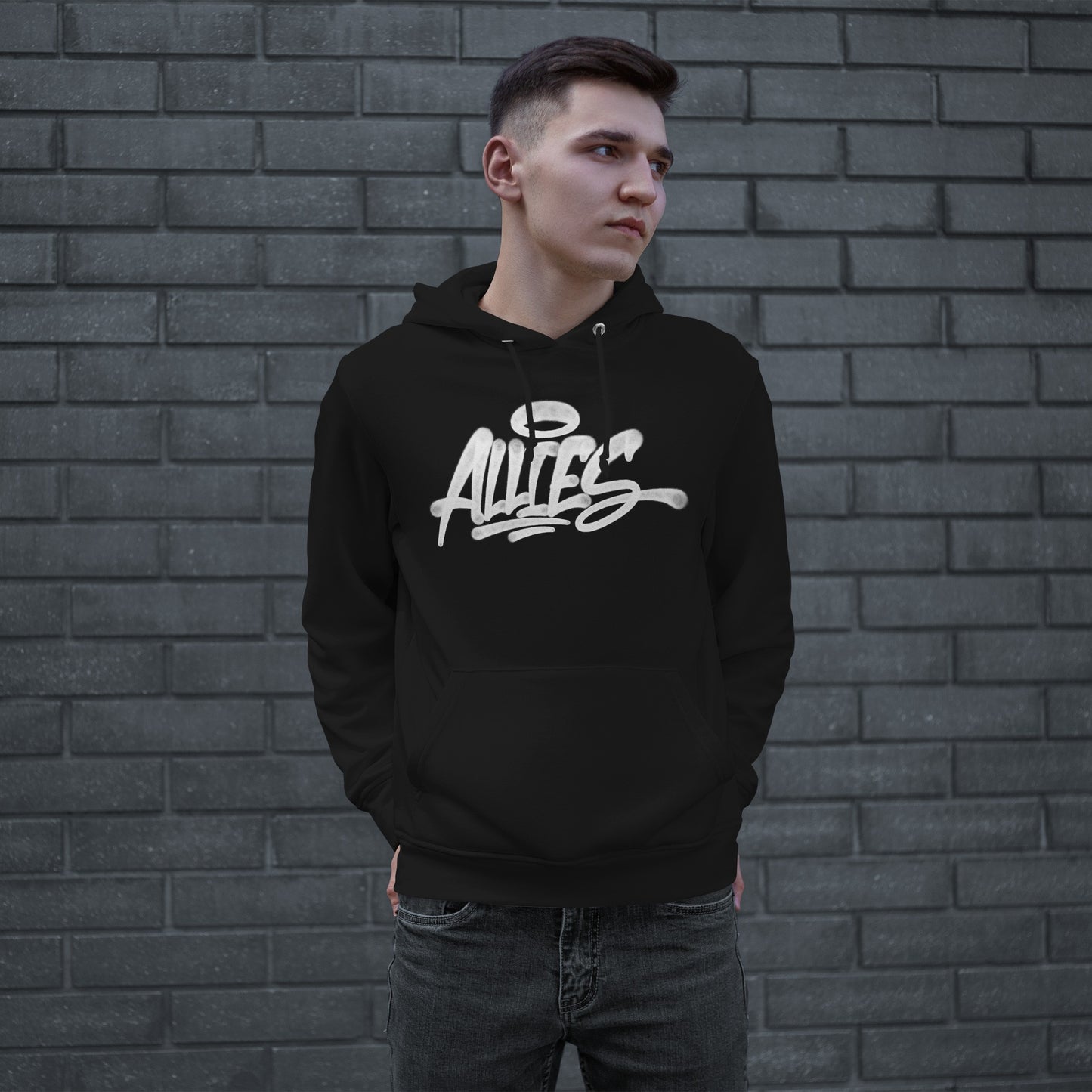 Young man wearing the Allies Handstyle Hoodie, featuring bold, graffiti-style script across the chest, embodying a statement of allyship and comfort, against a gray brick wall.