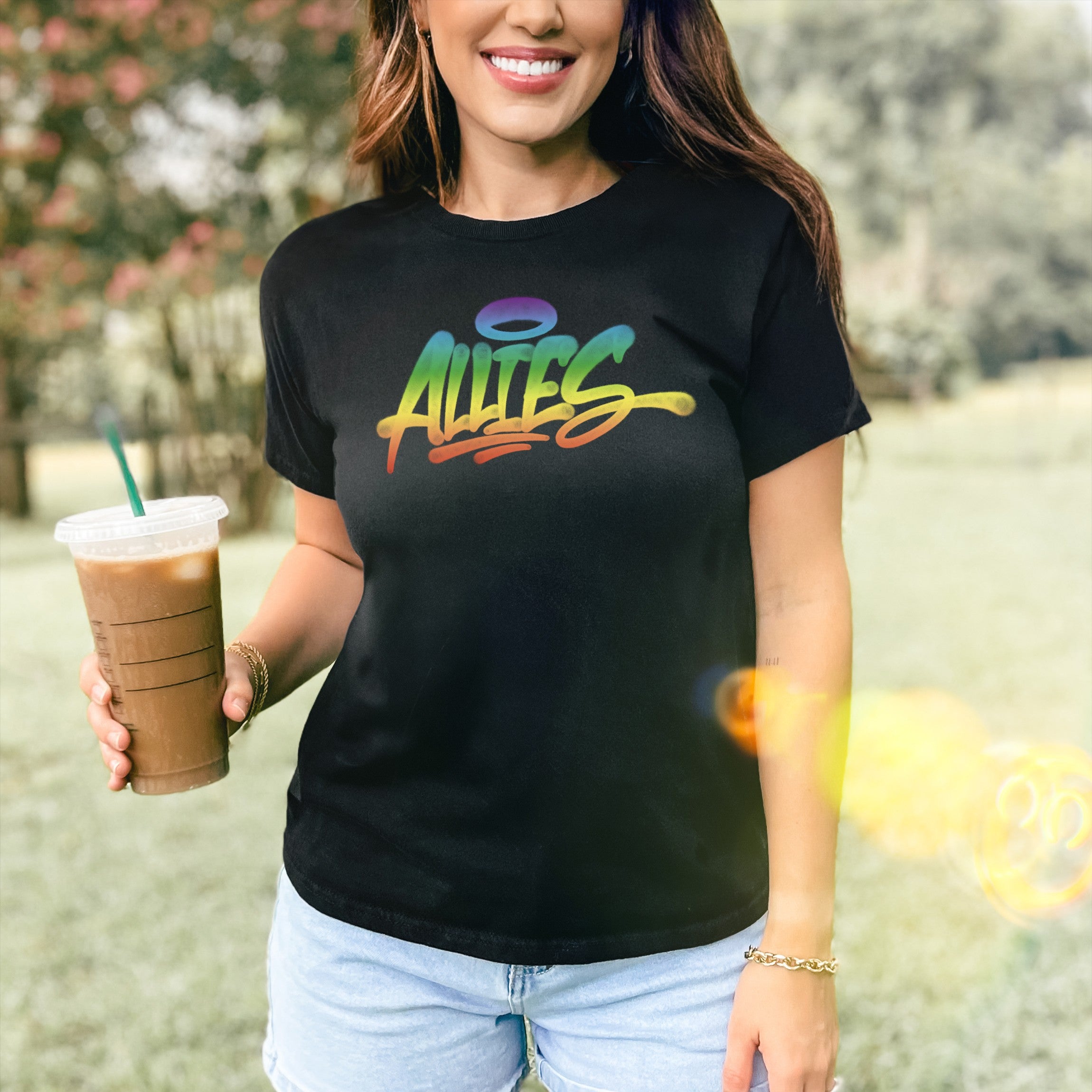 Person wearing the Allies Rainbow Handstyle Women's Baby Tee, featuring a rainbow-gradient Allies script with a halo motif, holding iced coffee outdoors. Tee embodies LGBTQ+ solidarity and pride.