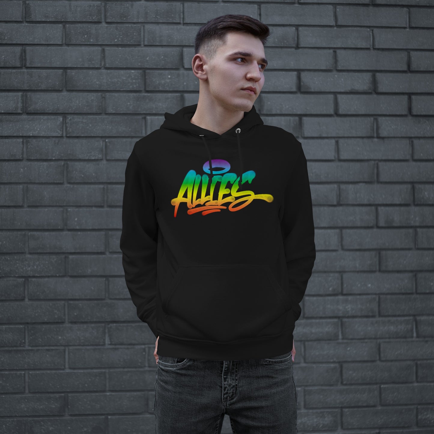 Young man in a black Allies Rainbow Handstyle Hoodie, featuring vibrant rainbow script with a halo, stands casually against a gray brick wall, embodying boldness and solidarity in urban street-style fashion.
