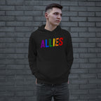 Young man in black Allies Rainbow Hoodie & Sweatshirt with rainbow-colored ALLIES text, symbolizing pride and unity, stands against a dark gray brick wall, hands in pocket, gazing sideways.
