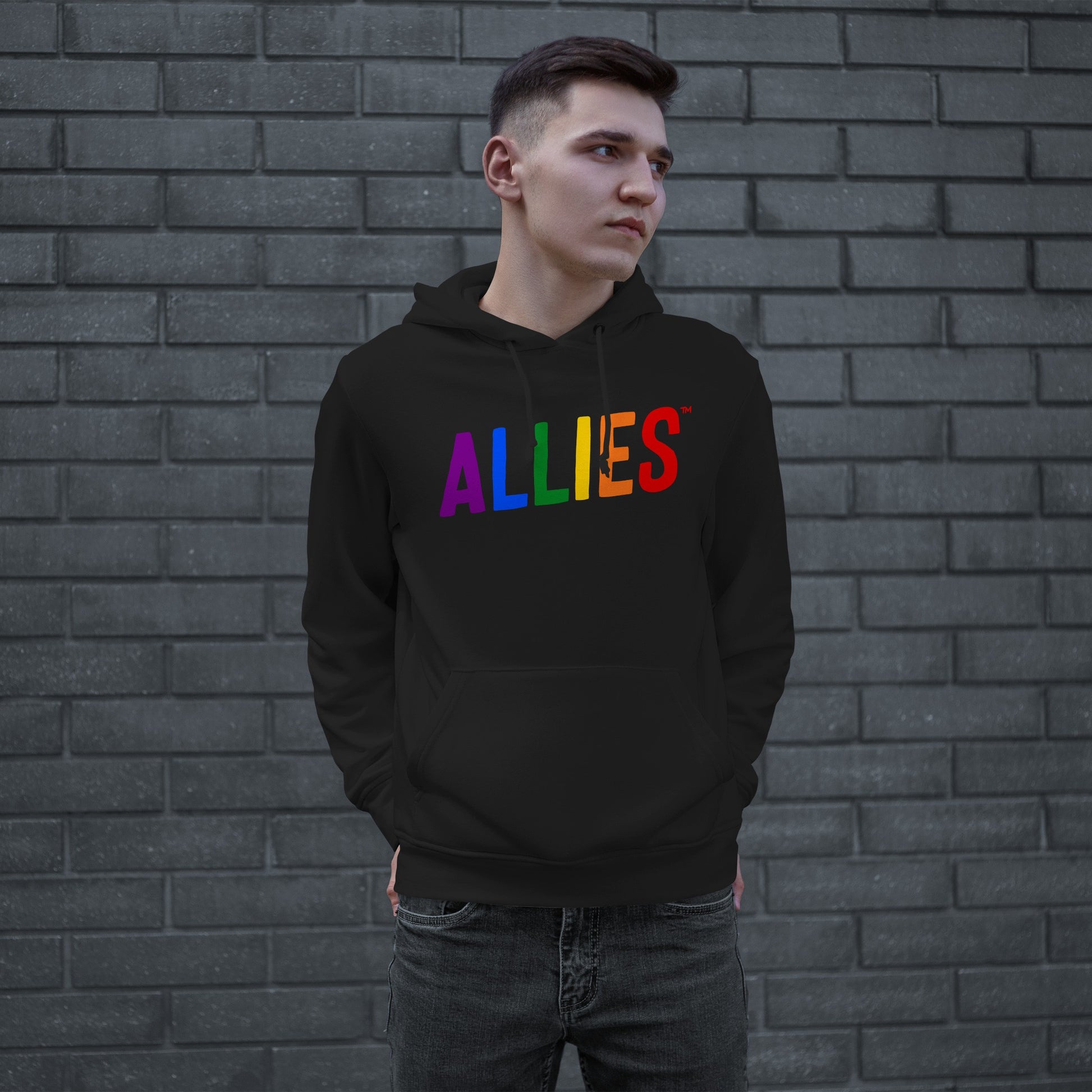 Young man in black Allies Rainbow Hoodie & Sweatshirt with rainbow-colored ALLIES text, symbolizing pride and unity, stands against a dark gray brick wall, hands in pocket, gazing sideways.