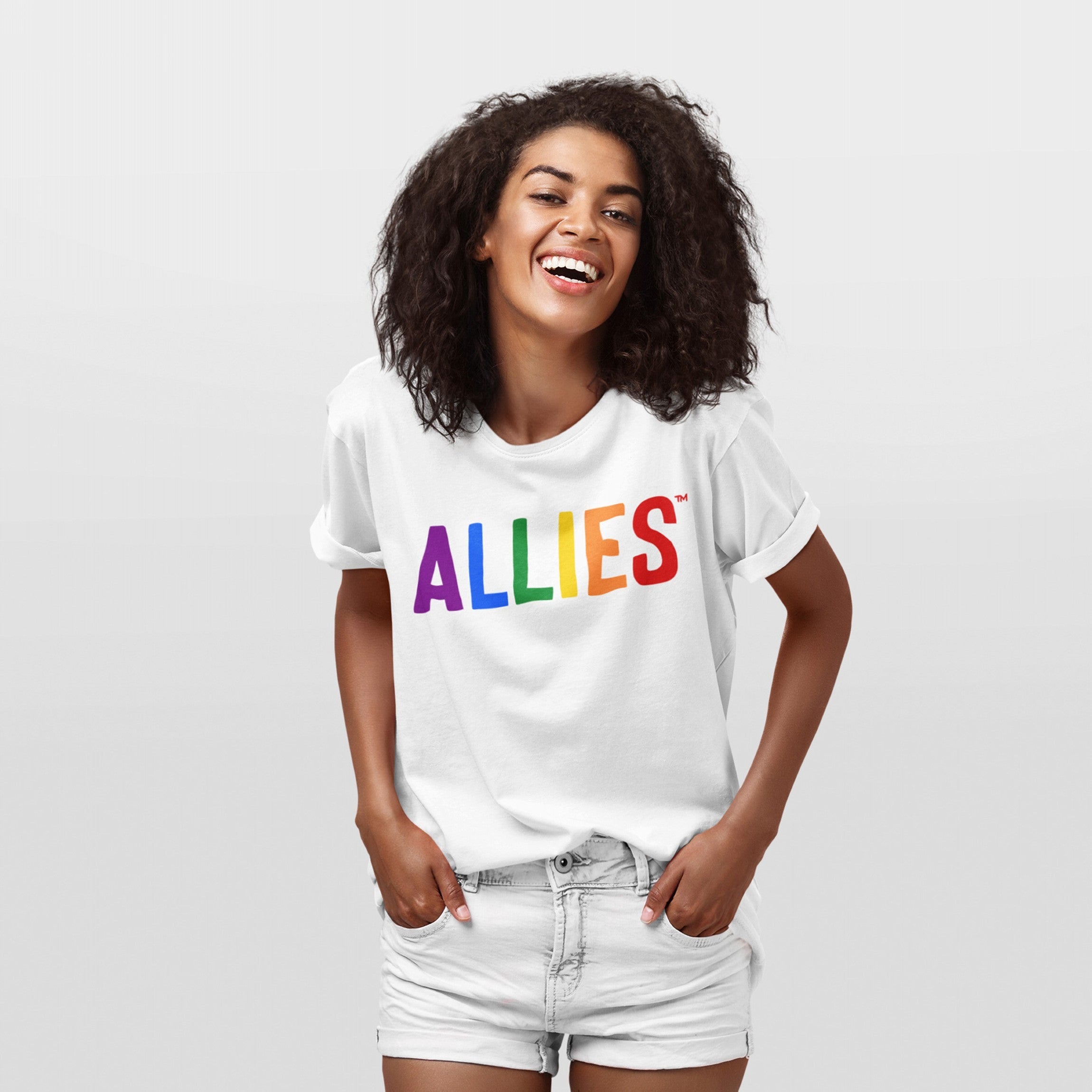 Young woman smiling in an Allies Rainbow T-shirt, featuring ALLIES in rainbow colors, embodying support for LGBTQ+ rights. She pairs it with light-wash denim shorts, hands in pockets.