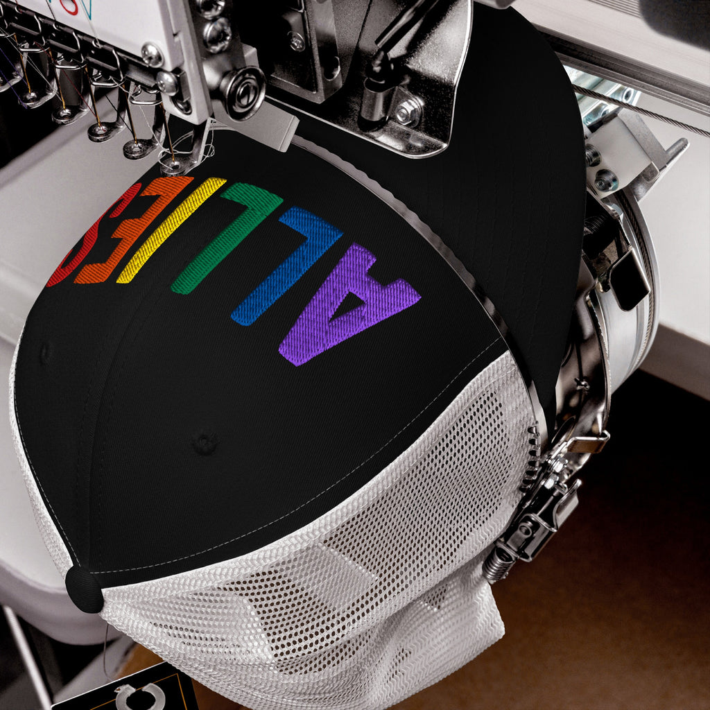 Automatic embroidery machine stitches ALLIES in rainbow gradient on a black mesh-back trucker hat, highlighting LGBTQ+ support. The hat is clamped, showcasing the vibrant, multi-colored letters across its crown.