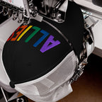 Automatic embroidery machine stitches ALLIES in rainbow gradient on a black mesh-back trucker hat, highlighting LGBTQ+ support. The hat is clamped, showcasing the vibrant, multi-colored letters across its crown.