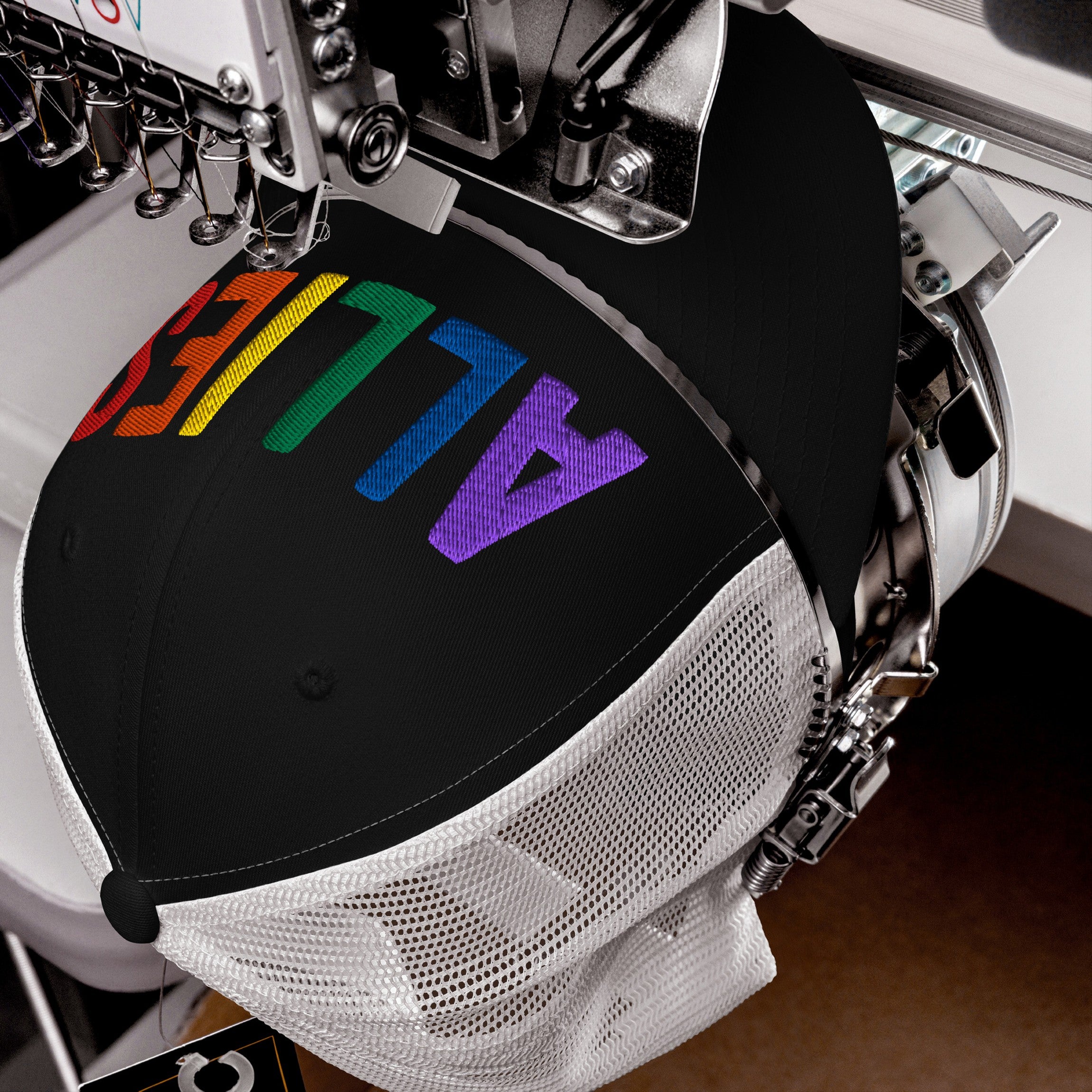 Automatic embroidery machine stitches ALLIES in rainbow gradient on a black mesh-back trucker hat, highlighting LGBTQ+ support. The hat is clamped, showcasing the vibrant, multi-colored letters across its crown.