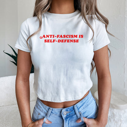 Person wearing the Anti-Fascism Is Self-Defense Women's Baby Tee, featuring bold red lettering on a fitted white crop-style T-shirt. Paired with light-wash jeans, embodying a stance of resistance and equality.