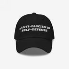 Black baseball-style cap with white embroidered text, “ANTI-FASCISM IS SELF-DEFENSE.” The Anti Fascism Is Self Defense Hat embodies activist spirit, ideal for protests or daily wear supporting human rights.