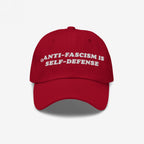 Red baseball cap with white embroidered text: ANTI-FASCISM IS SELF-DEFENSE. This Anti Fascism Is Self Defense Hat embodies bold activism, perfect for rallies or everyday resistance, supporting human rights and equality.