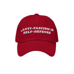 Red baseball cap with white embroidered text: ANTI-FASCISM IS SELF-DEFENSE. This Anti Fascism Is Self Defense Hat embodies bo