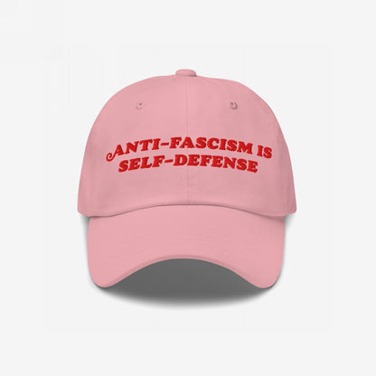 Light pink baseball cap with the slogan Anti-Fascism Is Self-Defense embroidered in red across the front, featuring a curved brim and classic six-panel design.