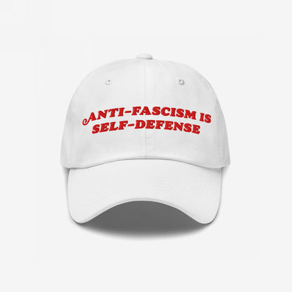 White baseball-style cap with red embroidered text ANTI-FASCISM IS SELF-DEFENSE, conveying a bold message of resistance. Perfect for advocates of equality and change, this durable, statement accessory supports Amnesty International.