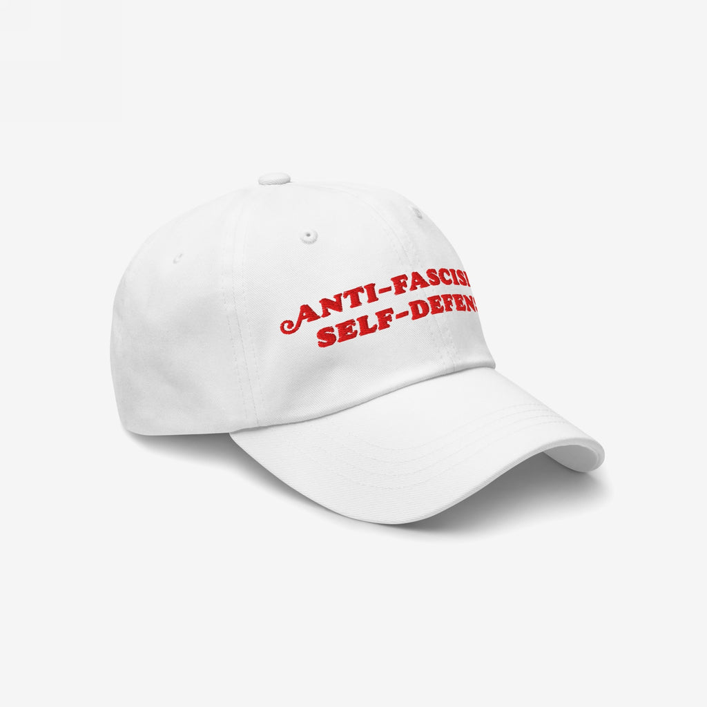 White baseball cap featuring bold red embroidery with the text ANTI-FASCISM and SELF-DEFENSE, embodying activist spirit and support for human rights causes. Ideal for protests and rallies.