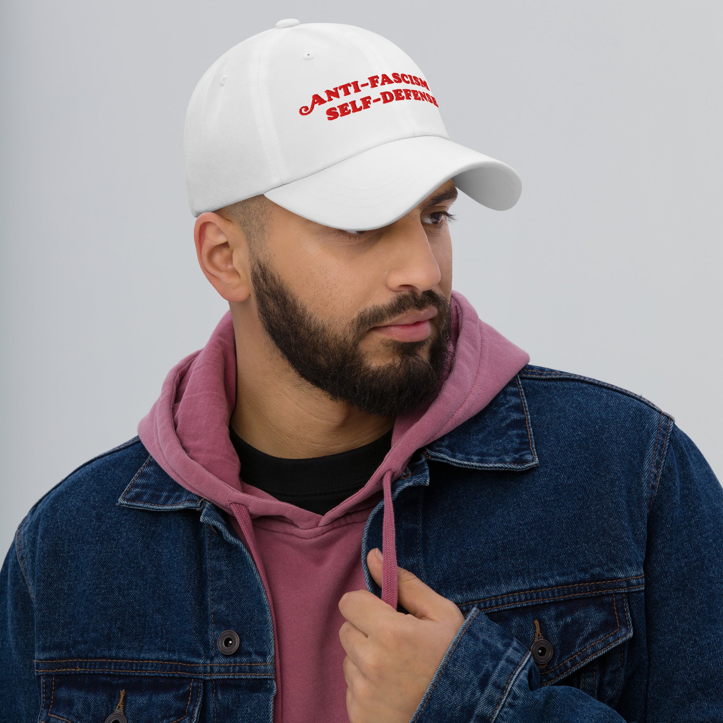 A bearded man models the Anti-Fascism Is Self-Defense Hat, featuring bold red embroidery, embodying activism and resistance. Ideal for advocates, it pairs a white cap with casual, layered attire.