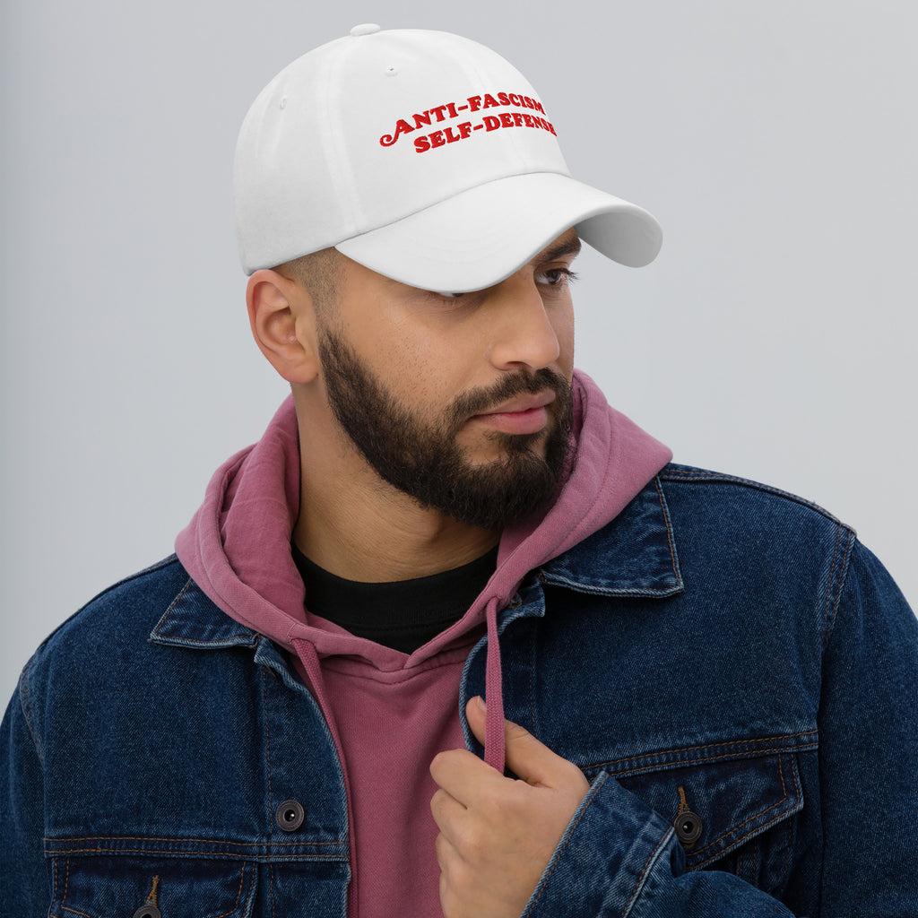 A bearded man models the Anti-Fascism Is Self-Defense Hat, featuring bold red embroidery, embodying activism and resistance. Ideal for advocates, it pairs a white cap with casual, layered attire.