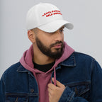 A bearded man models the Anti-Fascism Is Self-Defense Hat, featuring bold red embroidery, embodying activism and resistance. Ideal for advocates, it pairs a white cap with casual, layered attire.