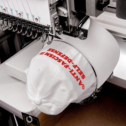 Multi-needle embroidery machine stitching “ANTI-FASCISM IS SELF-DEFENSE” on white cap, showcasing the Anti-Fascism Is Self-Defense Hat. Needles and thread guides visible, emphasizing resistance-themed fashion. Classic swing tag attached.