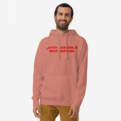 Smiling man models the Anti-Fascism Is Self-Defense Hoodie, featuring bold, impactful messaging on a muted red backdrop, embodying a stand against hate and promoting equality.