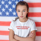 Young girl in a white Anti-Fascism Is Self-Defense T-shirt stands confidently, embodying activism and resistance, against a large American flag backdrop.