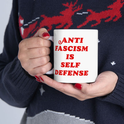 Person holding the Anti-Fascism Is Self-Defense Mug, featuring bold red text on a white ceramic surface. The mug is gripped with both hands, displaying red-painted fingernails and a dark sweater.