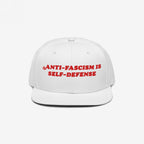 White flat-brimmed cap featuring bold red embroidery reading ANTI-FASCISM IS SELF-DEFENSE for activists and allies, embodying a vintage, unapologetic stance against fascism.