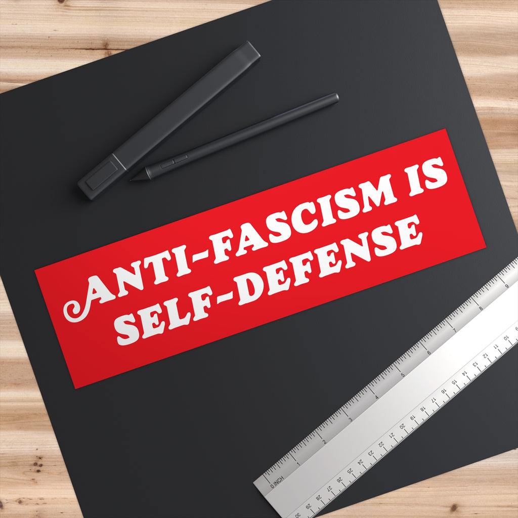 Anti Fascism Is Self Defense Sticker & Magnet on a dark tabletop, featuring bold white slogan on red. Nearby, a stylus pen with case and metal ruler, embodying a strong political statement.