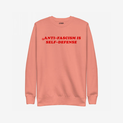 Coral-pink crewneck sweatshirt with bold red ANTI-FASCISM IS SELF-DEFENSE text across the chest, embodying the Anti-Fascism Is Self-Defense Hoodie & Sweatshirt's message of resistance and solidarity.