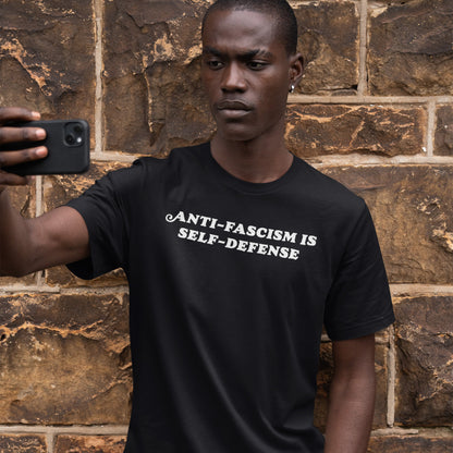 Young man holds a smartphone, wearing the Anti-Fascism Is Self-Defense T-shirt. The shirt features bold white lettering on black, embodying a clear, historical message of resistance and empowerment.