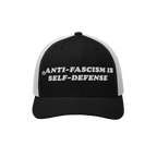 Two-tone trucker-style baseball cap with embroidered slogan “ANTI-FASCISM IS SELF-DEFENSE” on the front, embodying activi