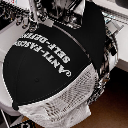 Black mesh-back trucker cap clamped in an embroidery machine, stitching Anti-Fascism Is Self-Defense in bold white script, embodying resistance.