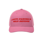 Pink trucker-style baseball cap with red embroidered “ANTI-FASCISM IS SELF-DEFENSE” text, embodying activist spirit. Idea