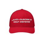 The image features the Anti Fascism Is Self Defense Hat, a red mesh-back trucker cap with ANTI-FASCISM IS SELF-DEFENSE embroi