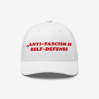 White mesh-back trucker hat with bold red embroidery reading “ANTI-FASCISM IS SELF-DEFENSE,” embodying activism and resistance. Ideal for allies and advocates, supporting ACLU's mission against fascism.