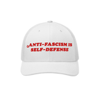 White mesh-back trucker hat with bold red embroidery reading “ANTI-FASCISM IS SELF-DEFENSE,” embodying activism and resis