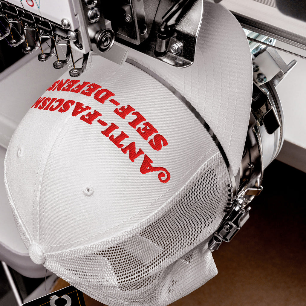 White mesh-back trucker cap on embroidery machine with red ANTI-FASCISM SELF-DEFENSE text stitched on front. Retro activist-style hat for protests, rallies, or everyday resistance.