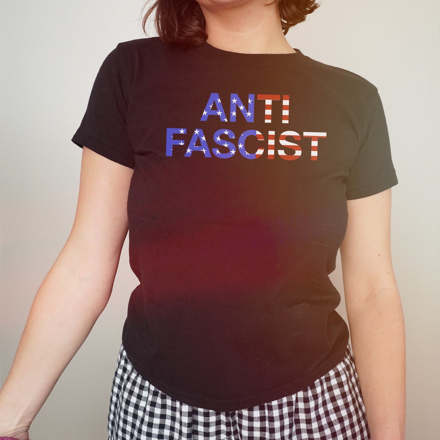 Person wearing the Anti-Fascist USA Classic Women's Baby Tee with an American flag pattern and black-and-white checkered bottoms, showcasing a bold statement against oppression and authoritarianism.