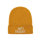 Anti Fascist Hat: A mustard-yellow, ribbed knit beanie with “ANTI FASCIST” embroidered in white on the cuff, embodying et