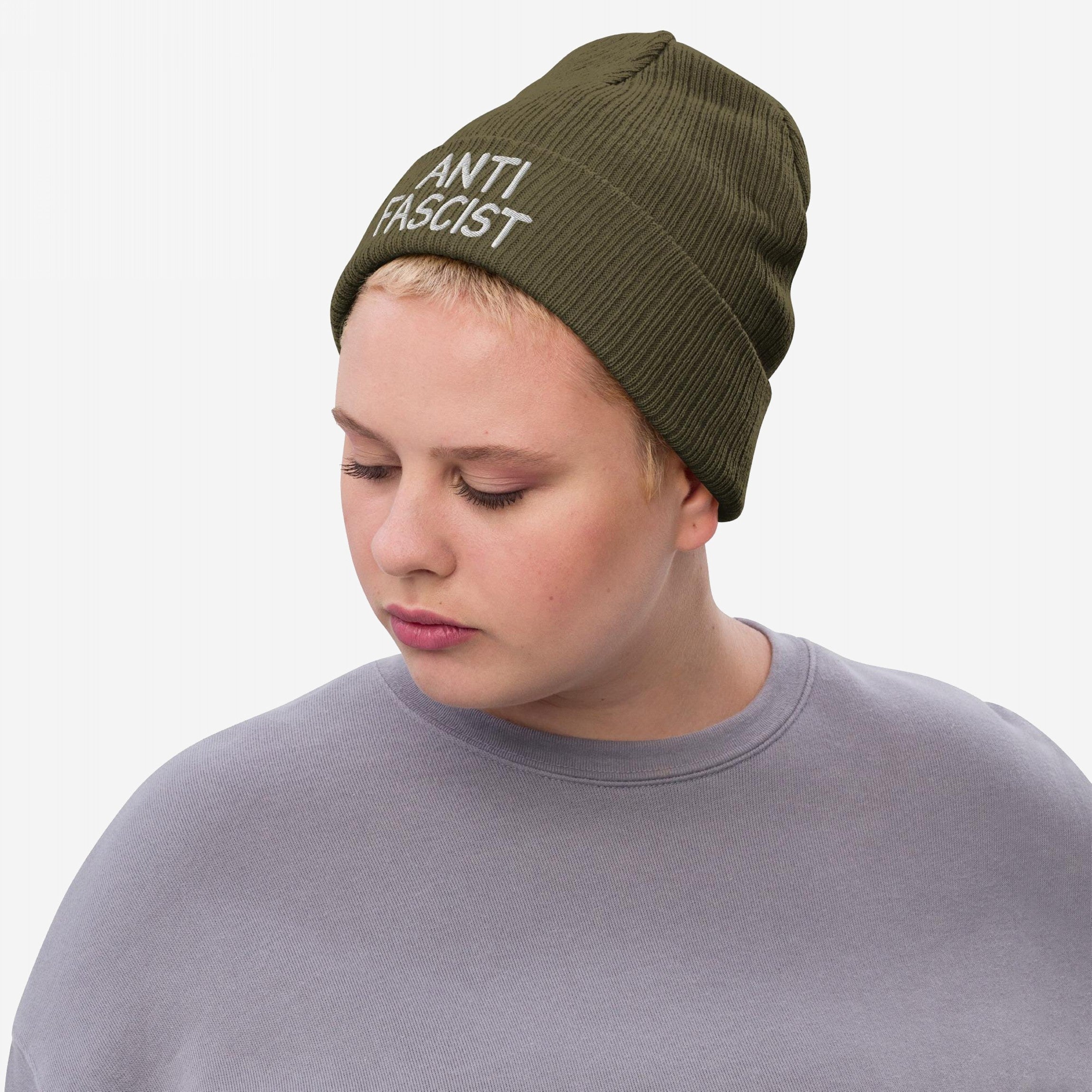 A person wearing the Anti Fascist Hat, an olive green rib-knit beanie with ANTI FASCIST embroidered in white, embodies defiance and style. Paired with a gray sweatshirt, it signifies bold activism.