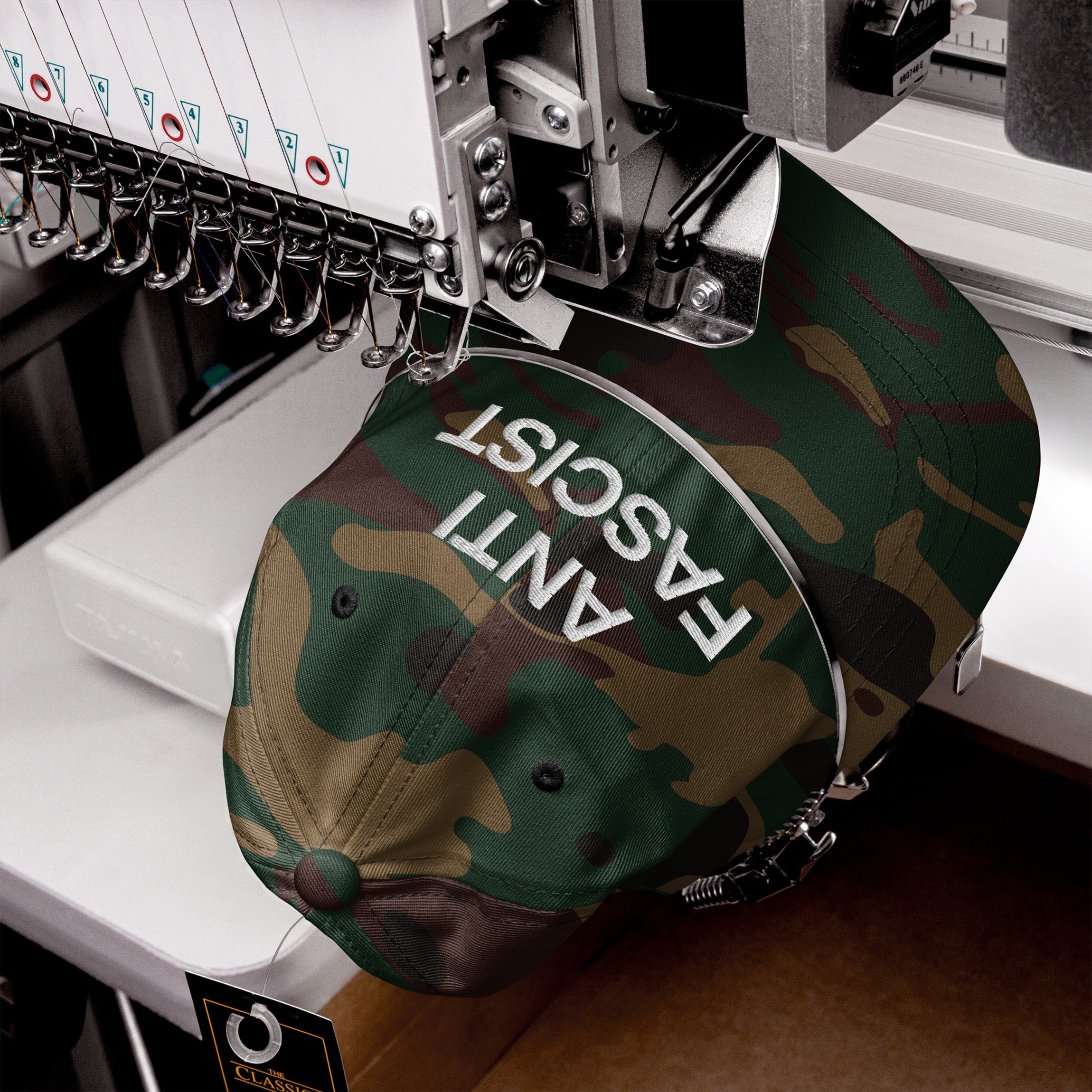 Anti-Fascist Classic Hat being embroidered with 'ANTI FASCIST' text on a camo-patterned baseball cap, showcasing bold resistance through sharp lettering on a multi-head embroidery machine.