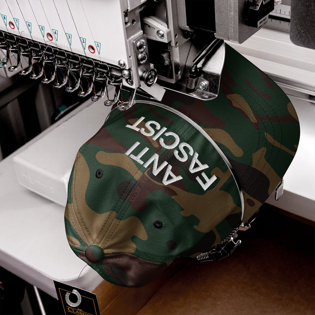 Anti-Fascist Classic Hat being embroidered with 'ANTI FASCIST' text on a camo-patterned baseball cap, showcasing bold resistance through sharp lettering on a multi-head embroidery machine.