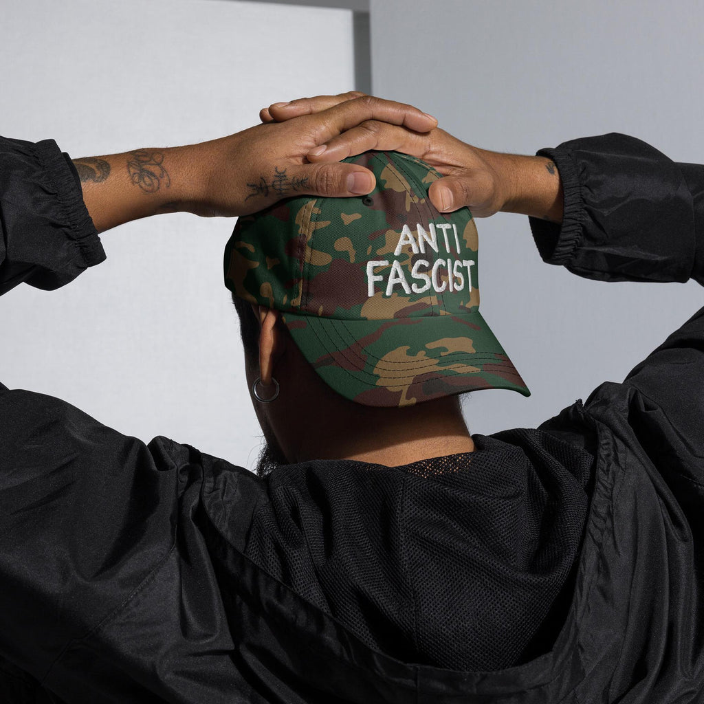 Person wearing the Anti Fascist Hat, featuring a camouflage pattern and ANTI FASCIST white embroidery, with tattooed arms raised to adjust the cap, embodying activism and style.