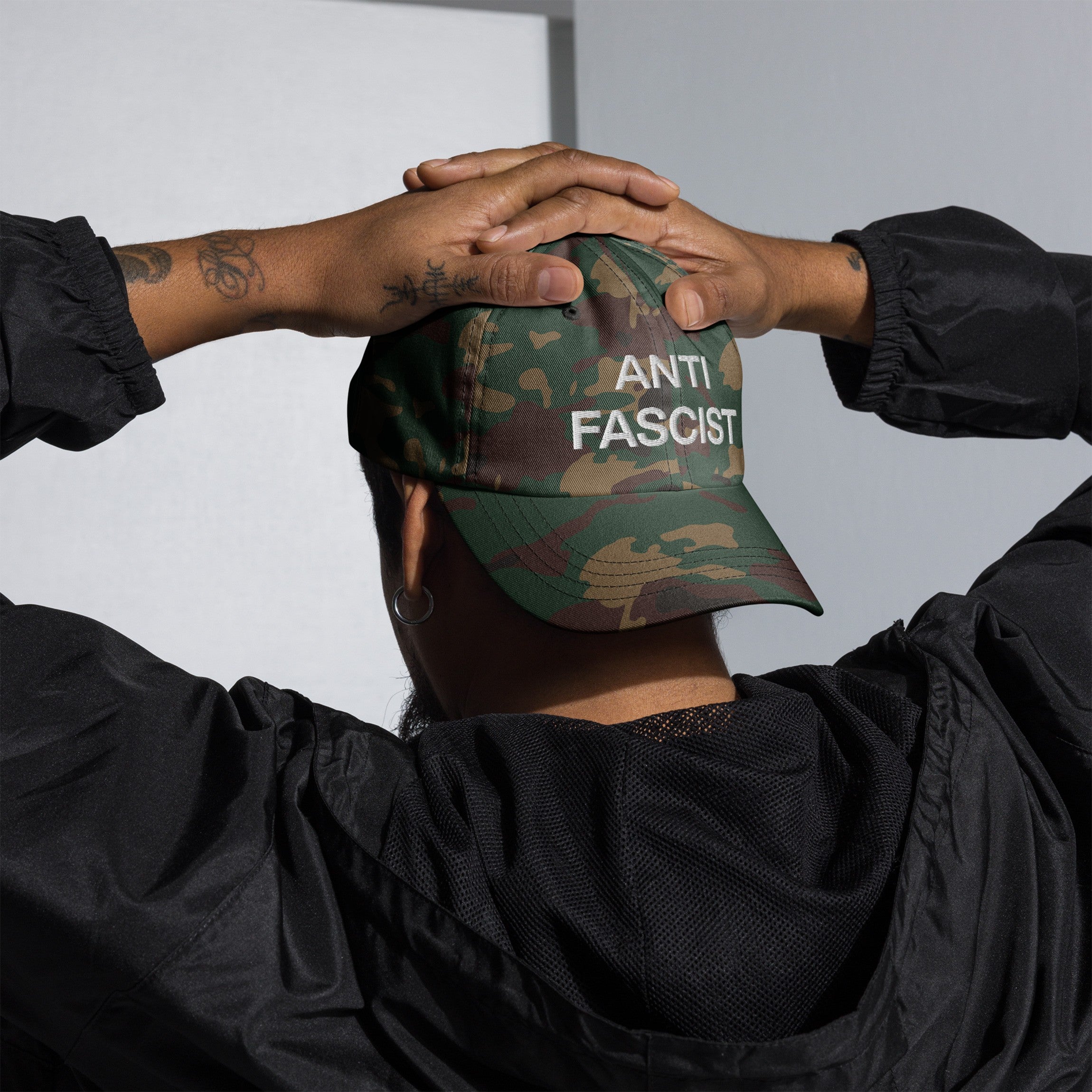 Person wearing the Anti-Fascist Classic Hat with camouflage print, embroidered with “ANTI FASCIST,” hands on head, showcasing a bold message of resistance through timeless style.