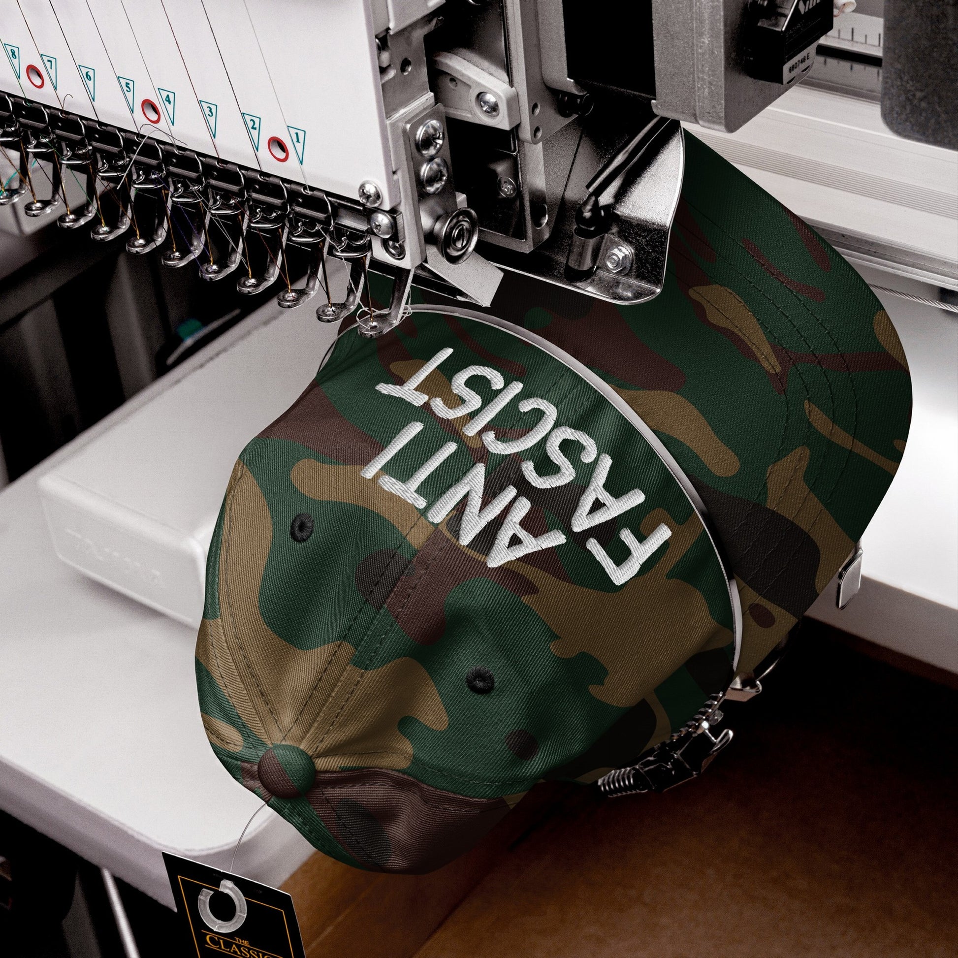 The Anti-Fascist Hat in a camouflage pattern is being embroidered with ANTI FASCIST in white on an industrial machine, featuring multiple needles and a Classic hang-tag on the bill.
