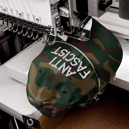 The Anti-Fascist Hat in a camouflage pattern is being embroidered with ANTI FASCIST in white on an industrial machine, featuring multiple needles and a Classic hang-tag on the bill.