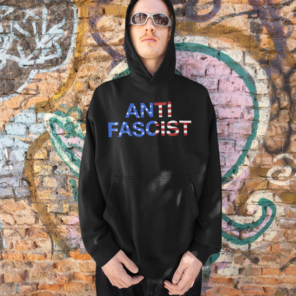 Young person in an Anti-Fascist USA Classic Hoodie & Sweatshirt with stars-and-stripes lettering, standing before a graffiti wall, embodying activism and resistance against tyranny.