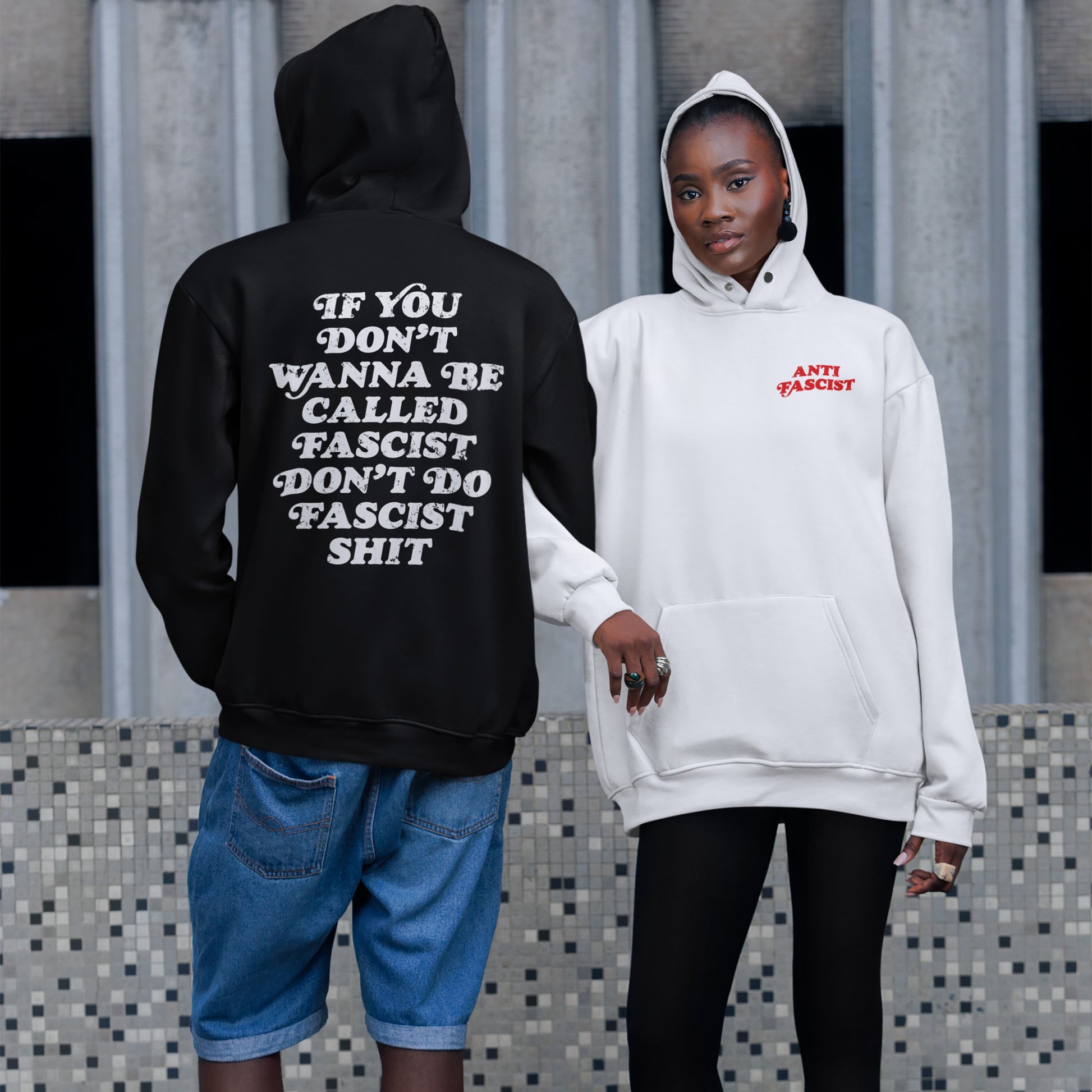 Two people showcase the Anti-Fascist Retro Shirt collection. One wears a black hoodie with a bold back message, while the other sports a light hoodie with ANTI FASCIST in red on the front.